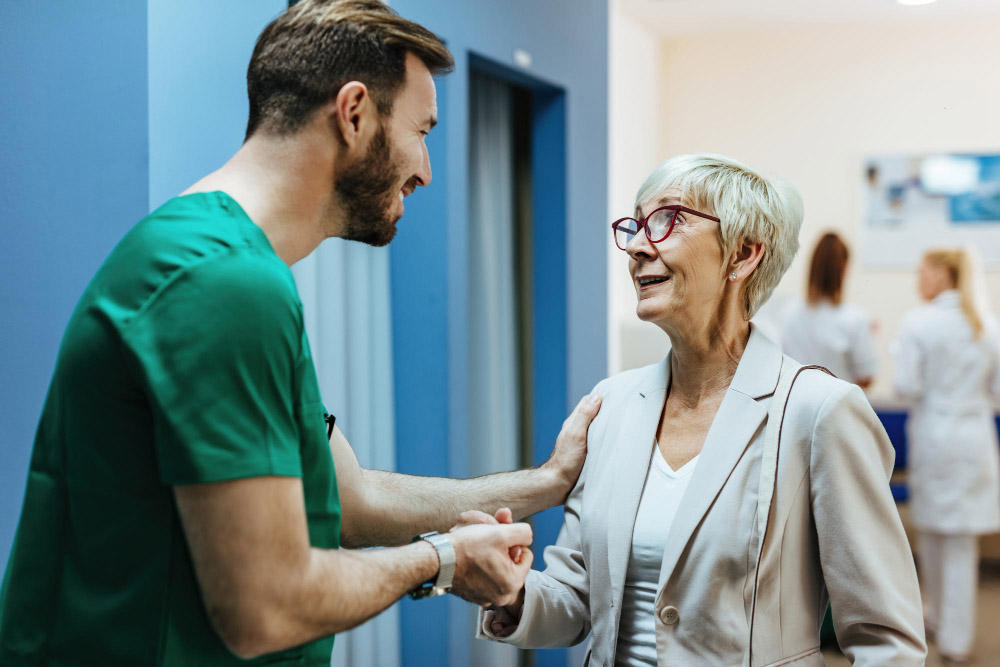 Pesquisa-de-Satisfacao-do-Paciente-mulher-senior-sorridente-de-maos-dadas-com-um-cirurgiao-enquanto-conversava-com-ele-em-um-corredor-do-hospital